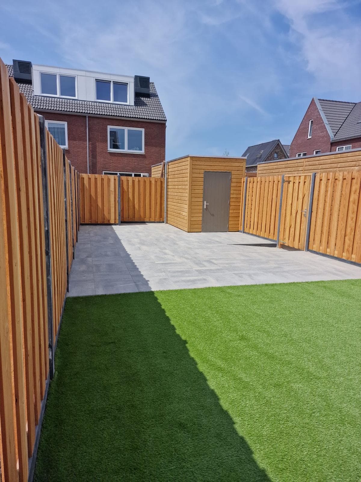 Aangelegde achtertuin met houten schuttingen, een tuinhuis, een bestraat terras en kunstgras