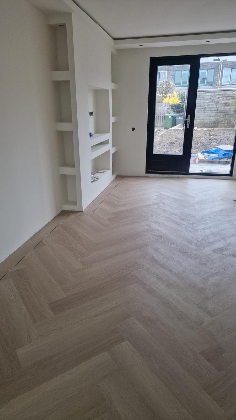 Lichte woonkamer met een nieuwe laminaatvloer in visgraatpatroon, een wand met nissen en tuindeuren
